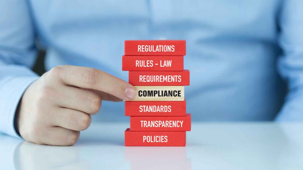 A hand pulling a _COMPLIANCE_ block from a stack of regulatory terms