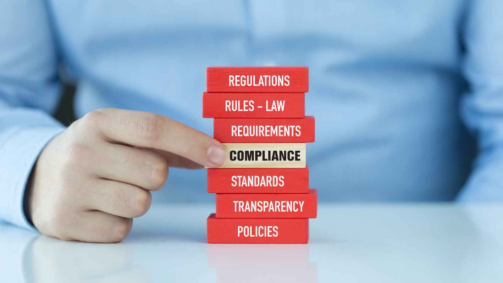 A hand pulling a _COMPLIANCE_ block from a stack of regulatory terms