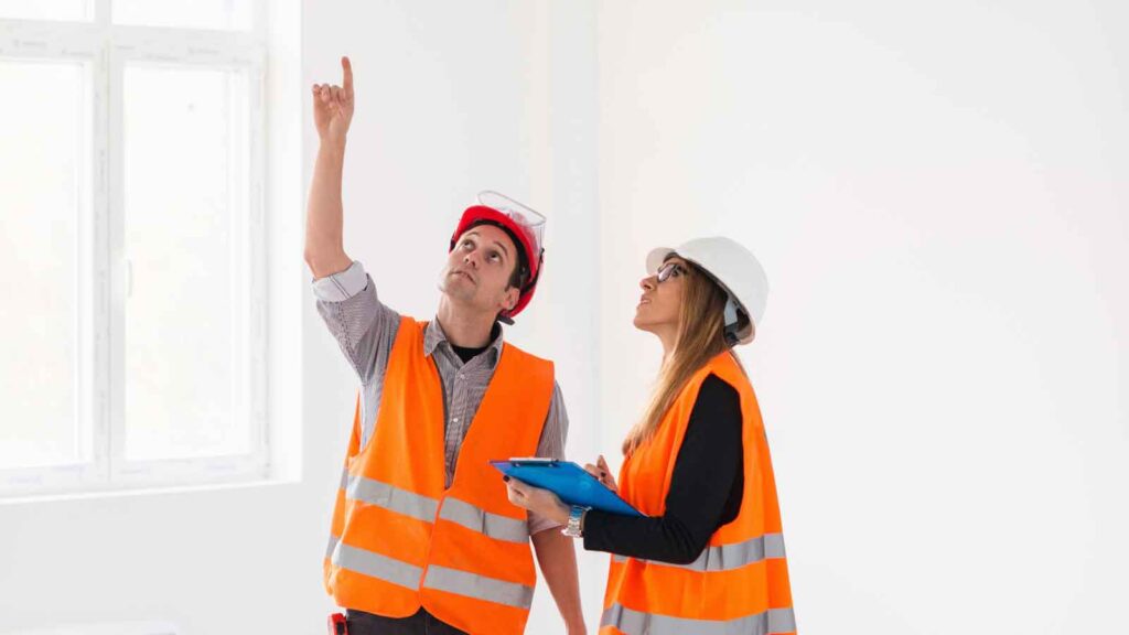 Two construction workers inspecting a ceiling in a bright room.